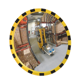 A round convex warehouse safety mirror with a yellow and black striped protective frame, reflecting a forklift and pallets inside a warehouse.